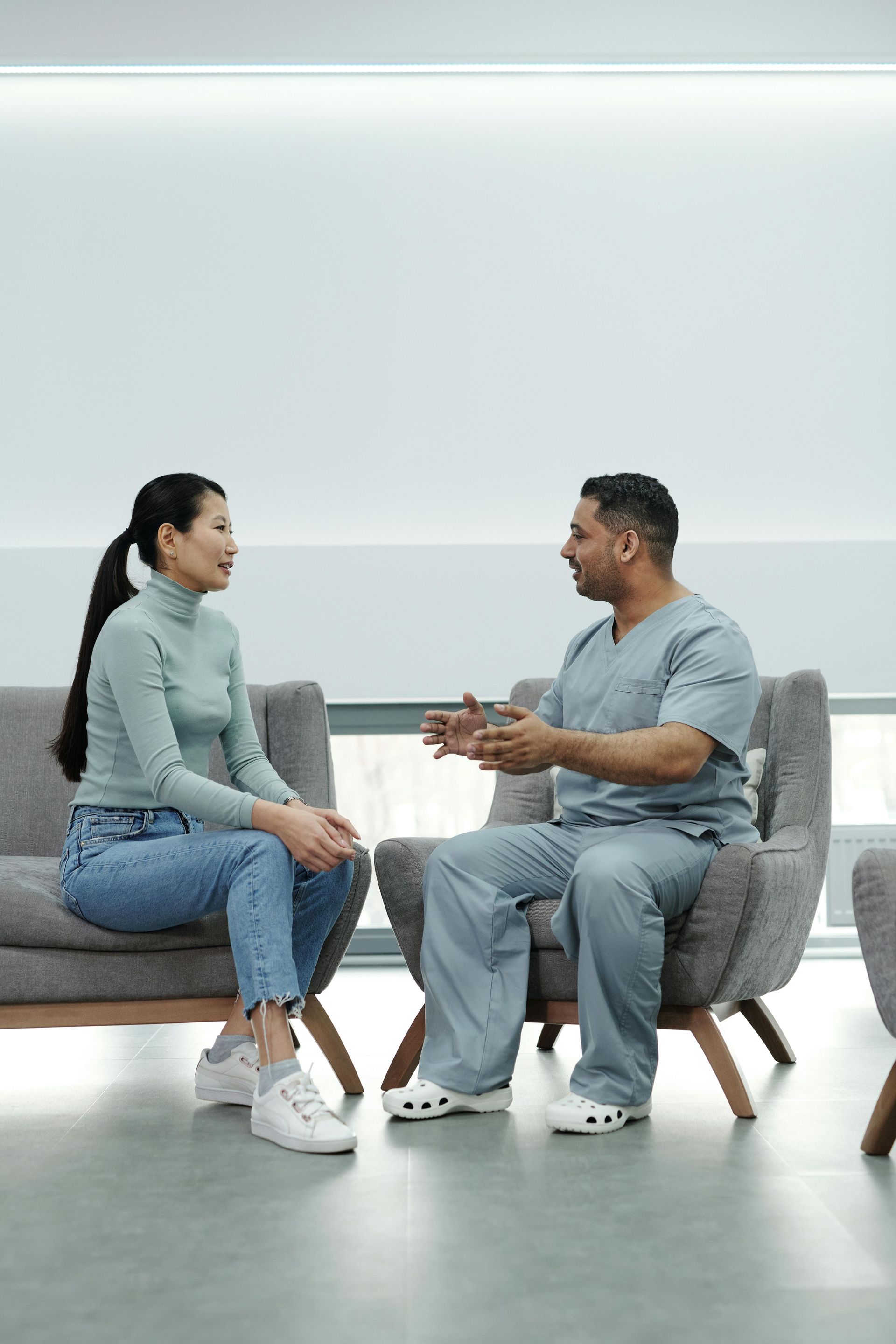 Woman in blue jeans and turtleneck speaks with a man in scrubs, seated in chairs in a neutral-lit room.