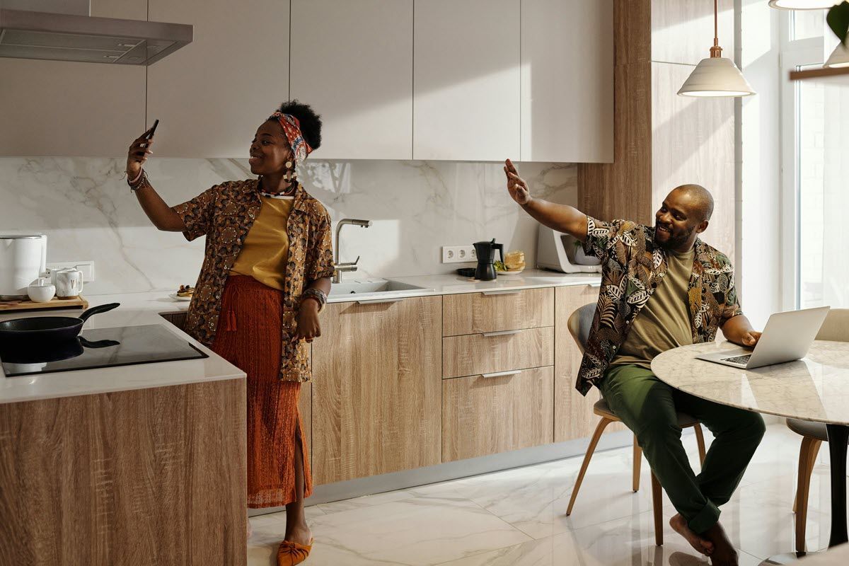 Woman taking selfie in kitchen while man waves at laptop, both wear patterned shirts.