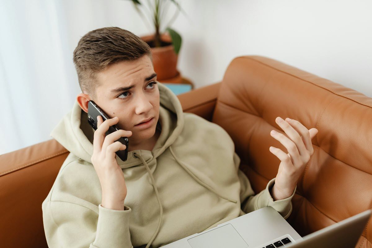 Teen on a couch using a phone and laptop, looking concerned, gesturing with a hand.