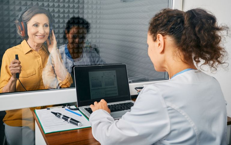 Woman in soundproof booth undergoing hearing test; audiologist at computer.