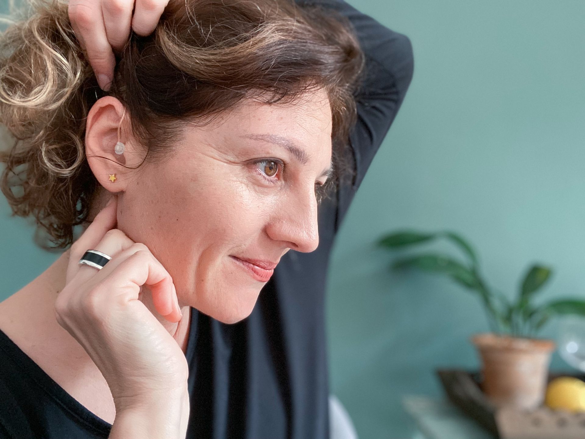 Woman in black shirt touching her hair, looking down with a slight smile; plant in background.