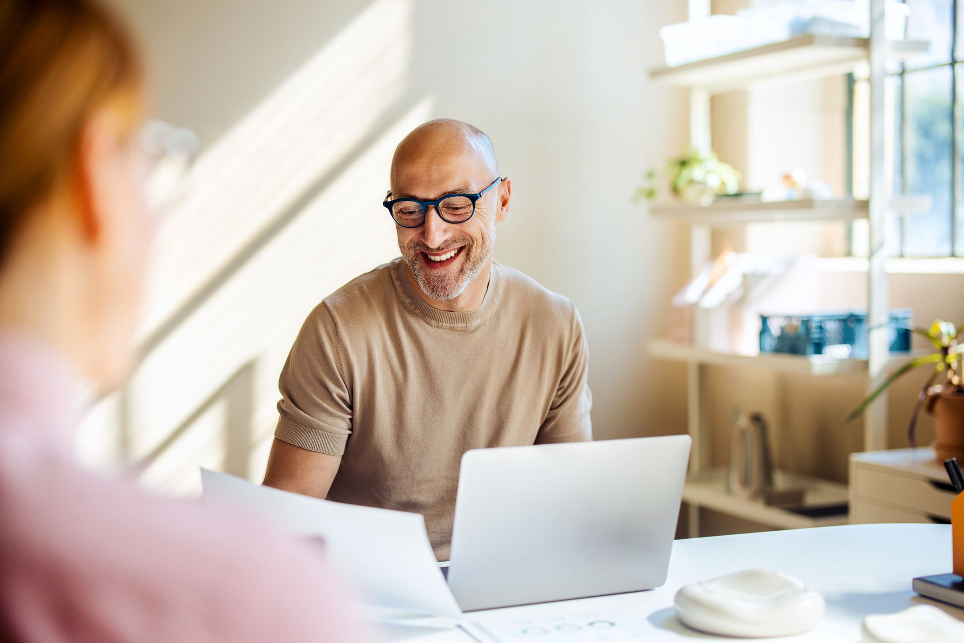 Man with glasses smiles, looking at a laptop and documents at a desk near a window.