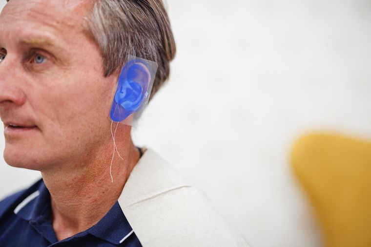 Man wearing a blue ear mold device with wires near his neck. White backdrop.