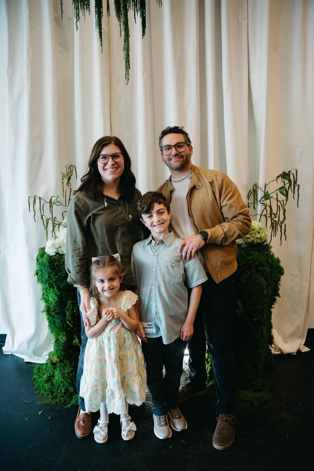 Family of four posing indoors in front of white curtains and greenery, smiling at the camera.