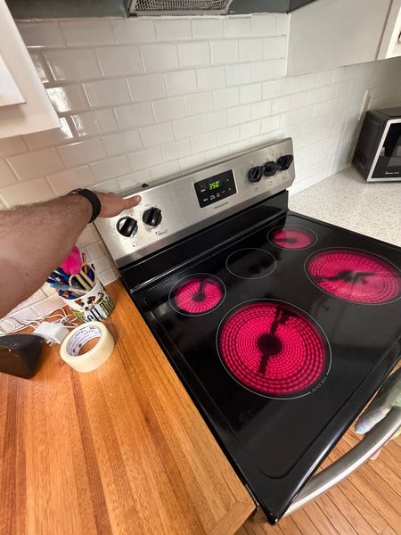 Hand on a stainless electric stove with glowing red burners in a kitchen.