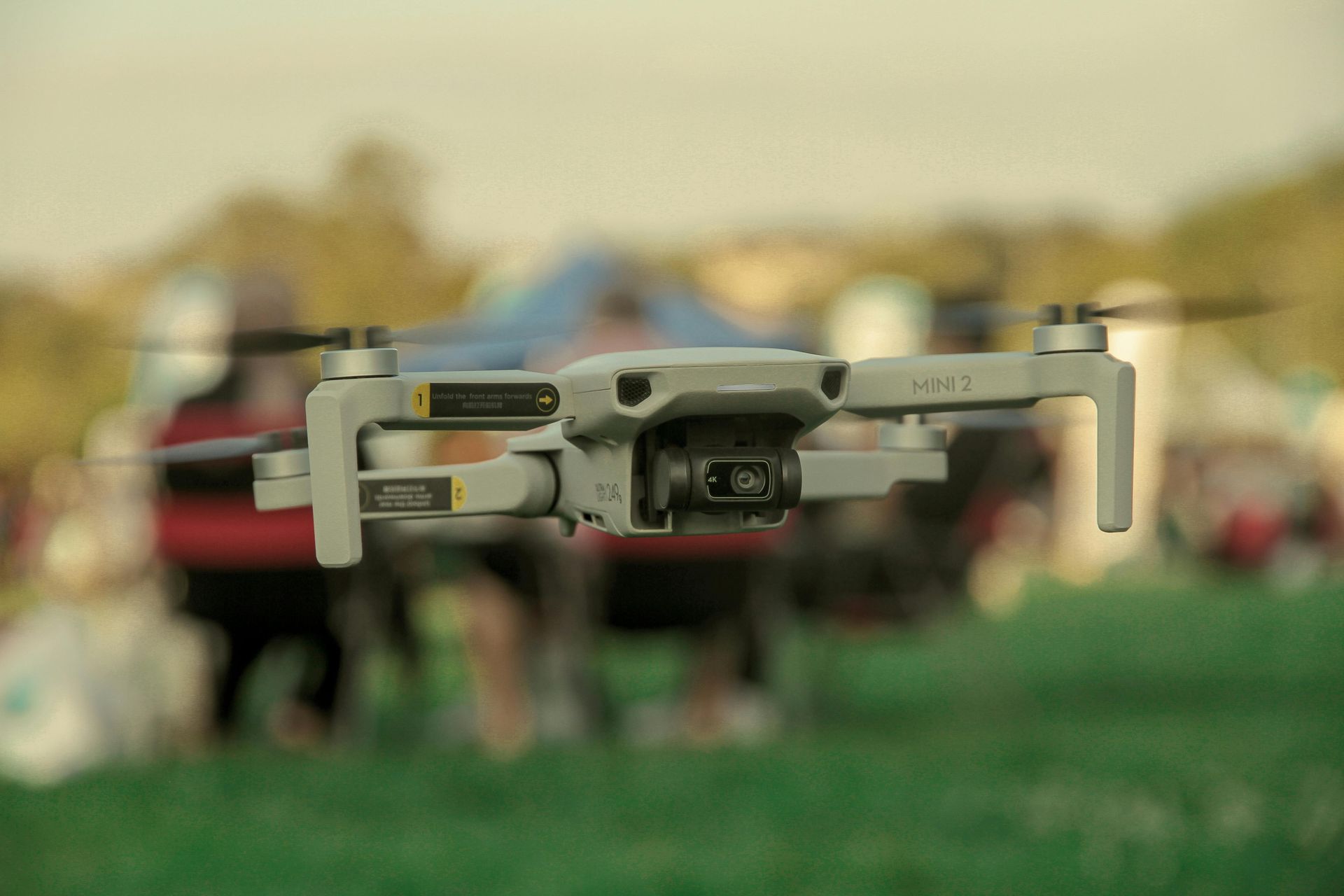 Drone hovering above a grassy field with people and a red vehicle blurred in the background