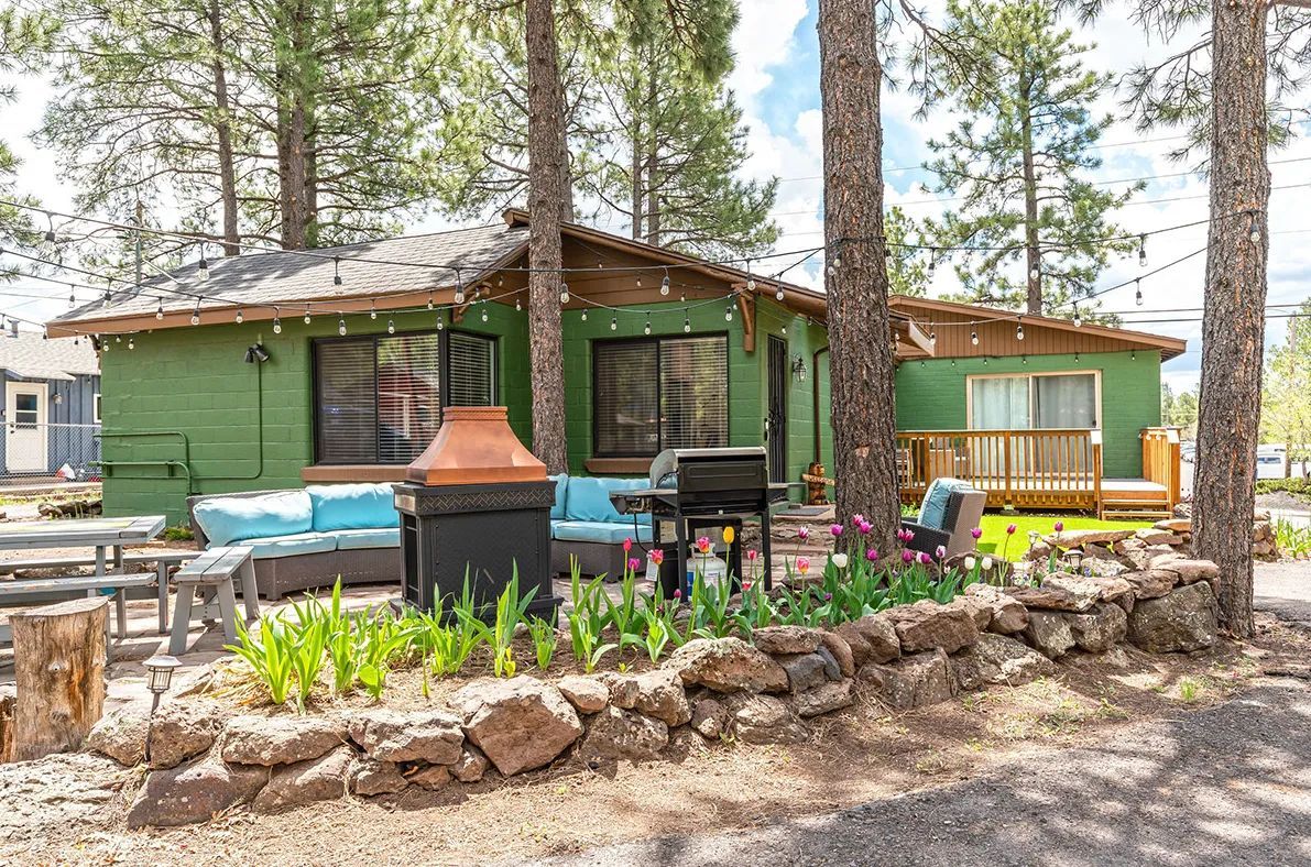 Green cabin with outdoor seating, grill, and rock garden, surrounded by trees.