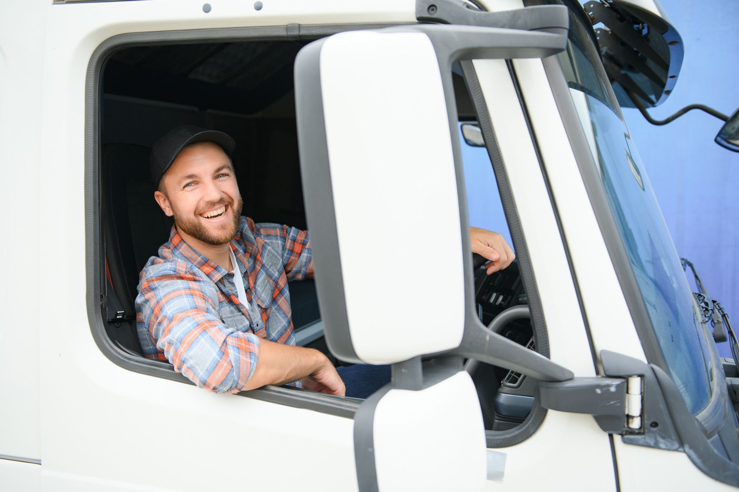 Smiling man in plaid shirt leaning out of a white truck window, waving on the road