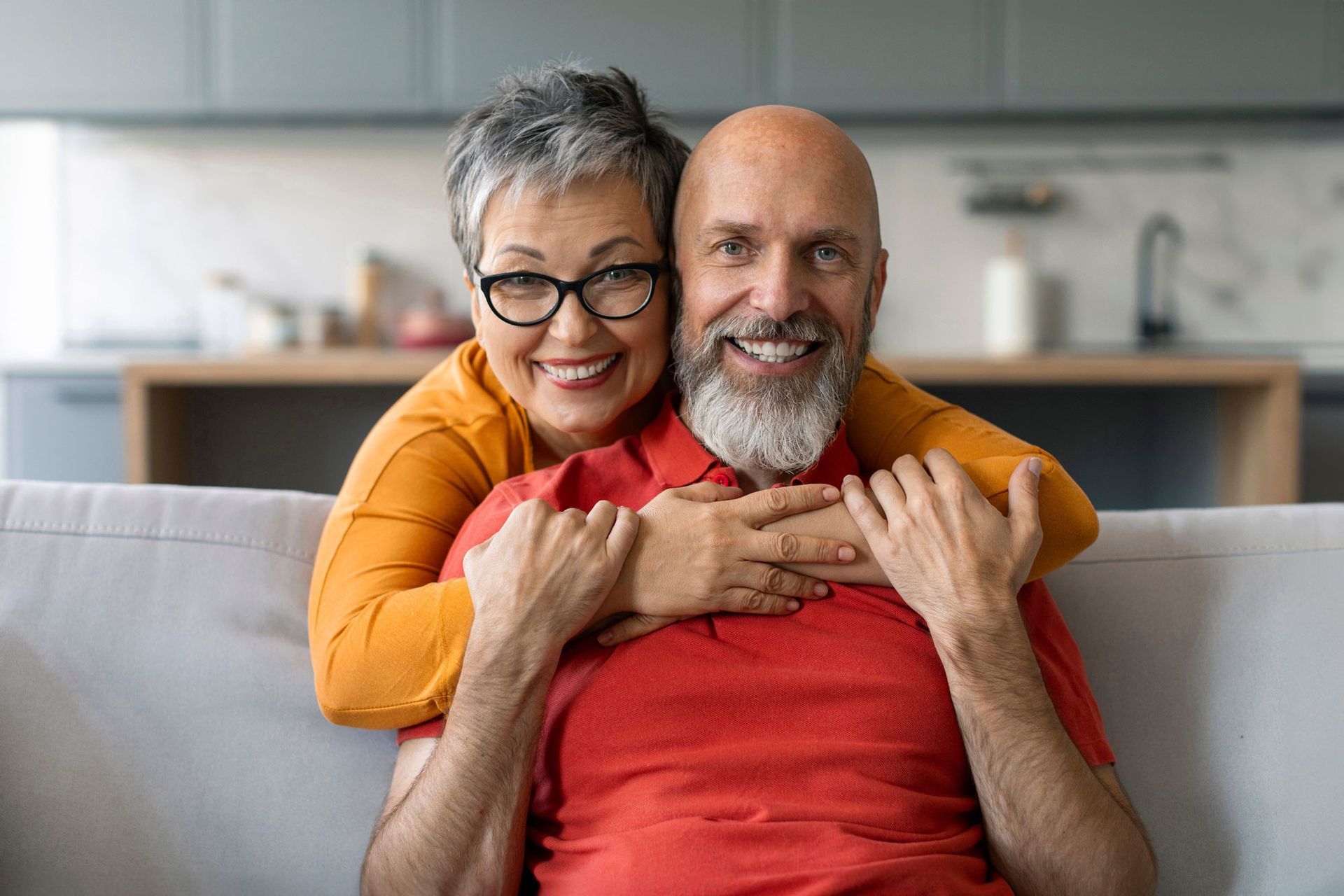 Smiling couple on a couch, with the woman hugging the man from behind in a bright living room.