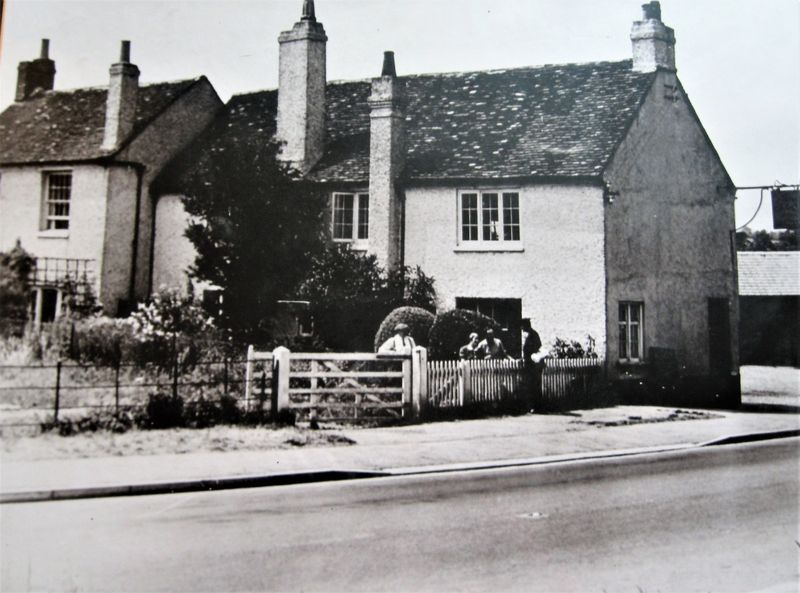 Black and white photo of two-story white cottages with picket fence and road in front.