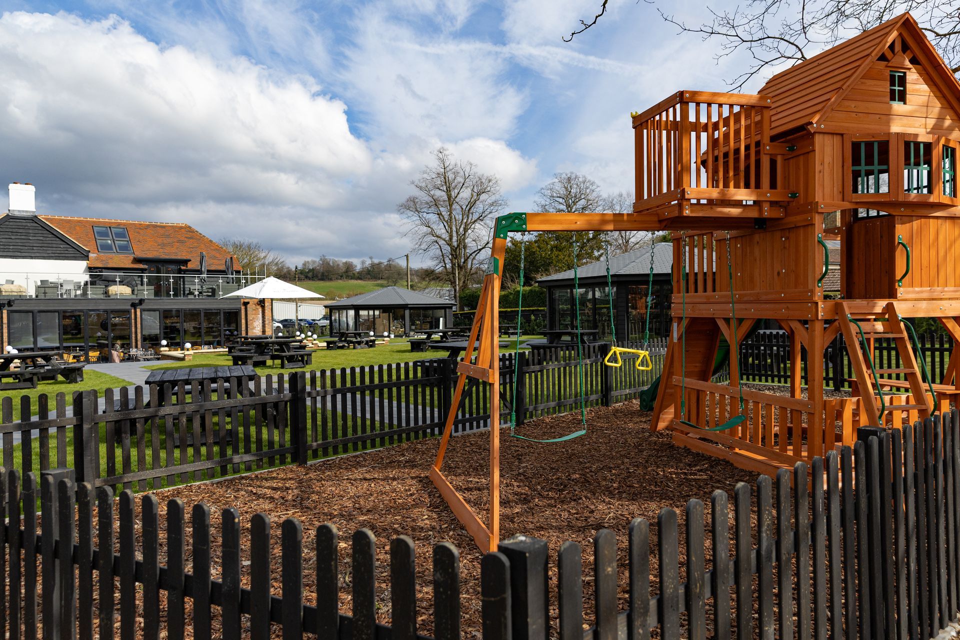 Playground with swings and a wooden playhouse in a pub garden on a cloudy day.