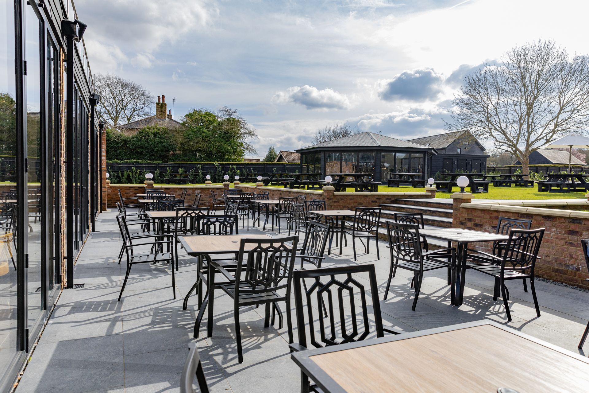 Outdoor patio with black tables and chairs, adjacent to a glass-walled structure, sunny day, gazebo in the distance.