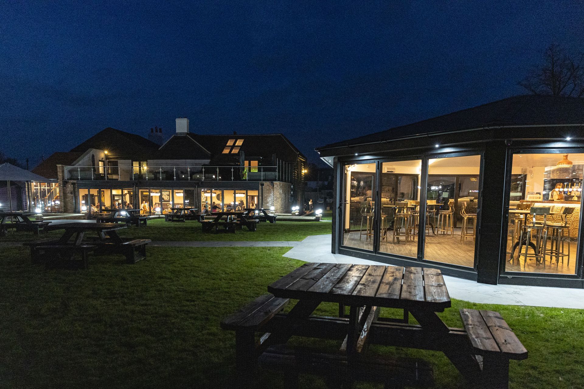 Nighttime view of a pub with outdoor seating. A gazebo-like structure is next to a large building, both lit.