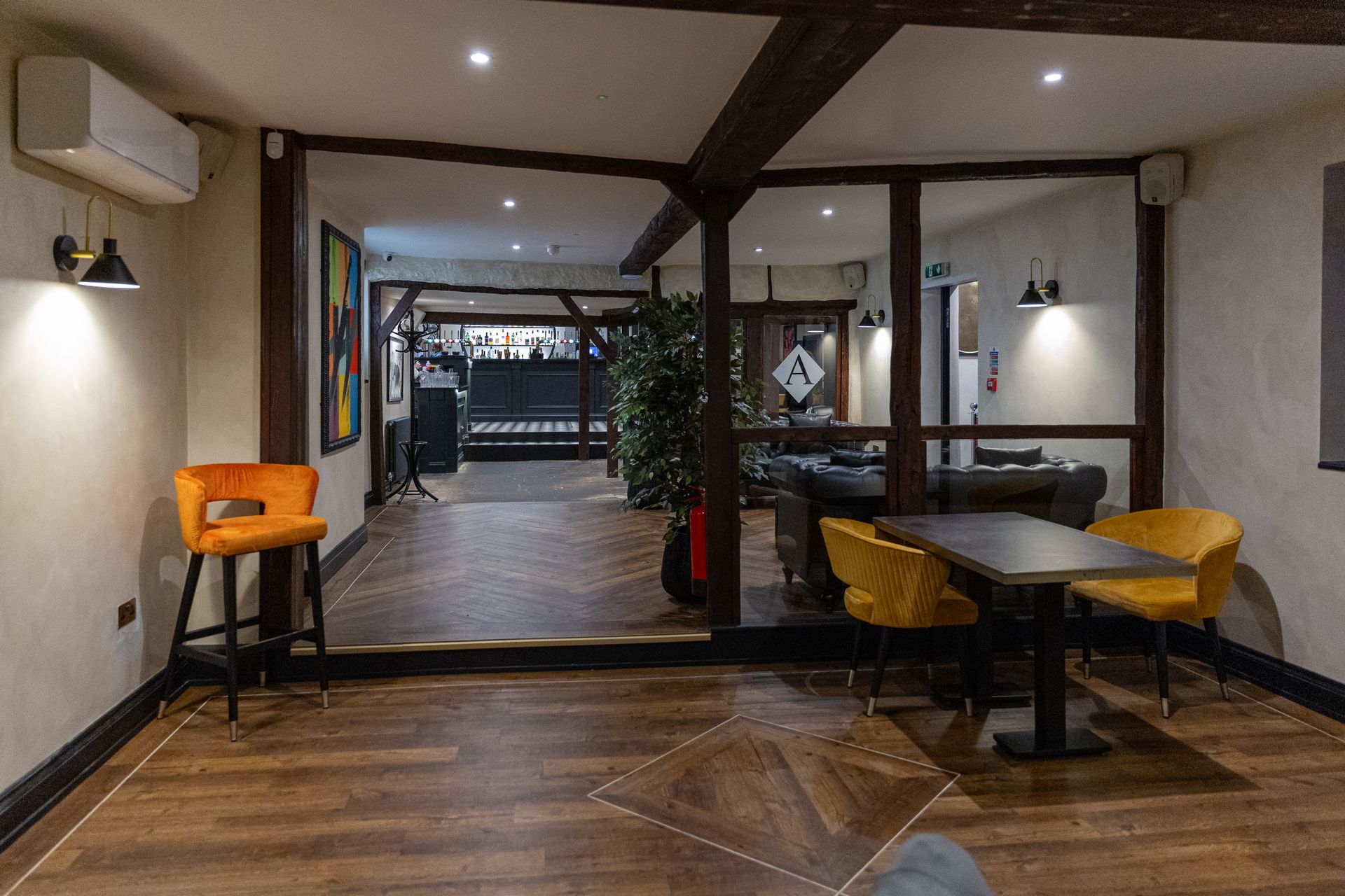 Interior with wooden beams, bar stools, small table with chairs, and a bar area in the background.