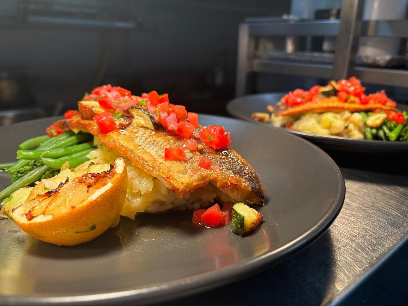 Pan-seared fish with tomato garnish, lemon, green beans, and zucchini on a gray plate.