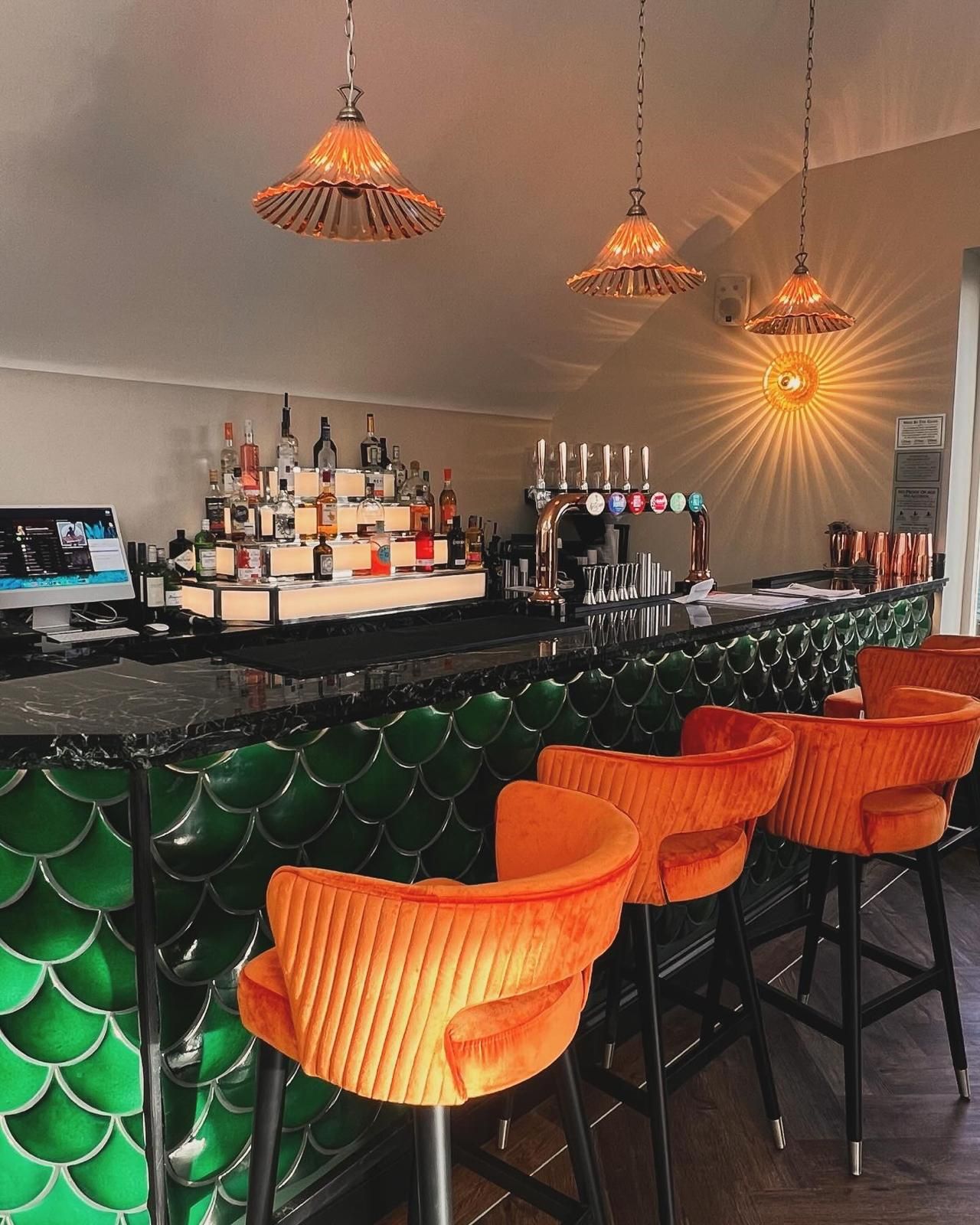 A bar interior with green tile, orange chairs, and hanging lights; liquor bottles on shelves.