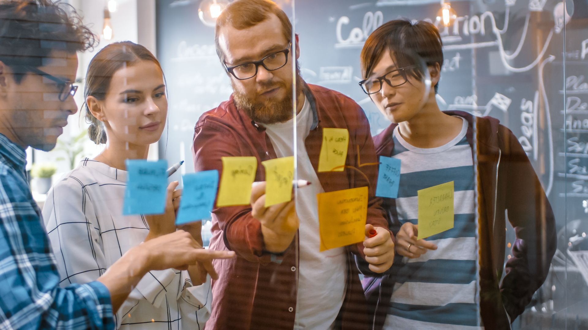 Group of people brainstorming, looking at sticky notes on glass. Indoors, natural light.