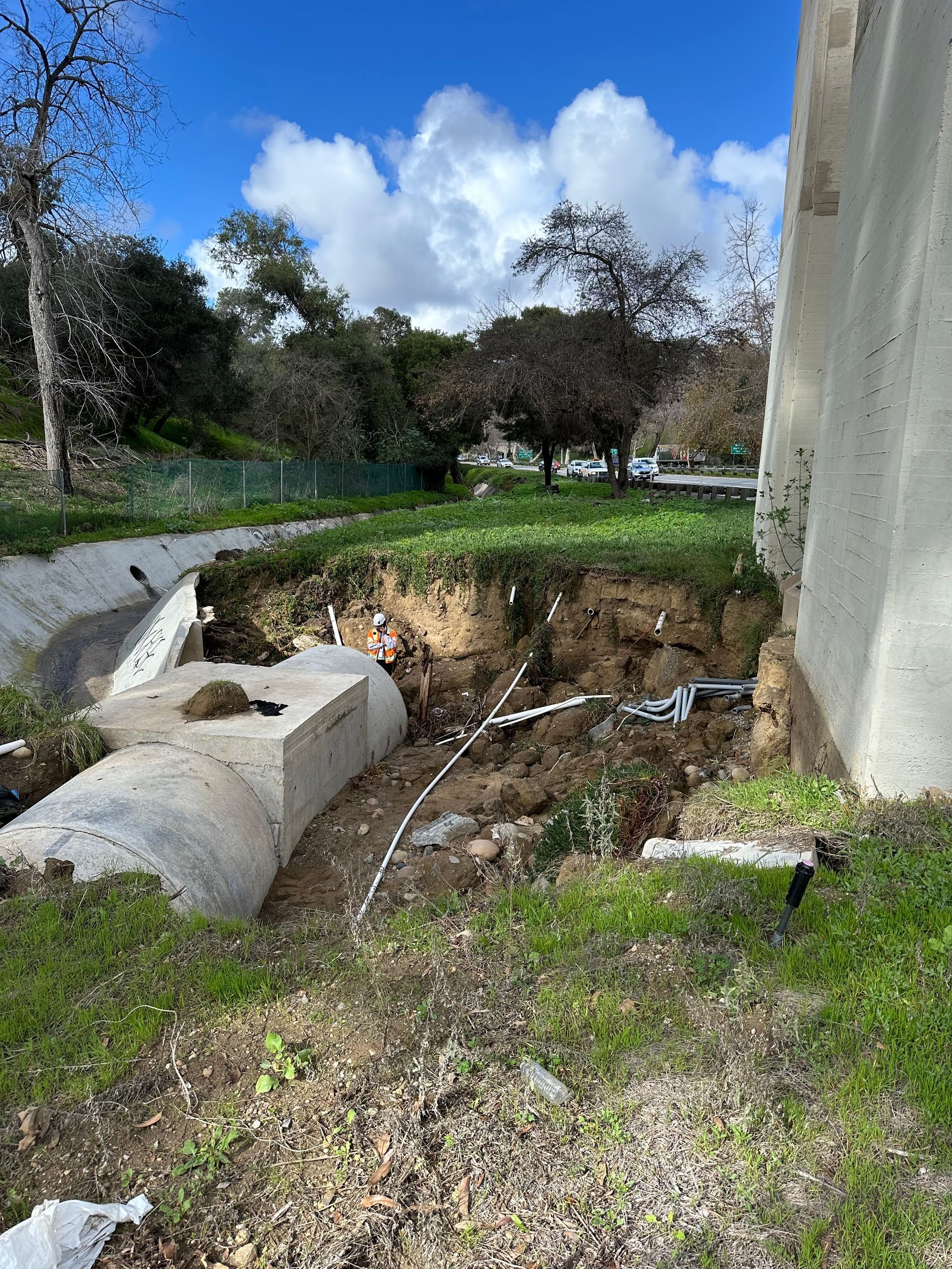 Erosion damage near concrete structure, exposed earth, grass, trees, and cloudy blue sky.