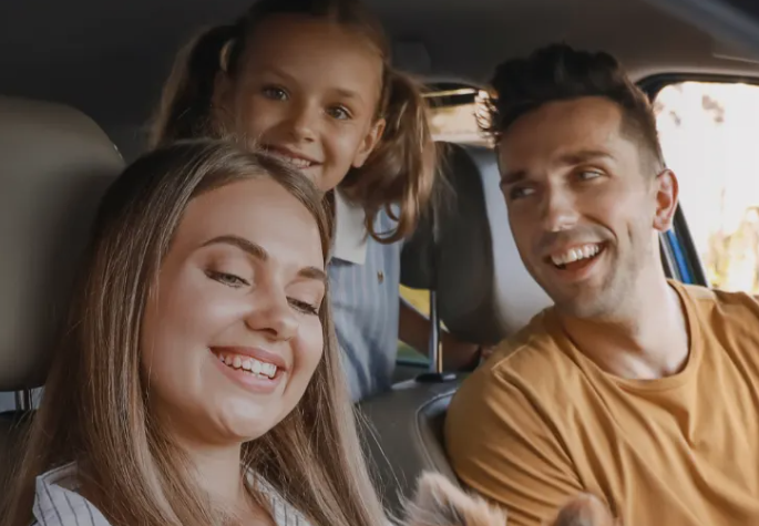 Família em um carro: pais sorrindo, criança com tranças espiando do banco de trás, dia ensolarado.