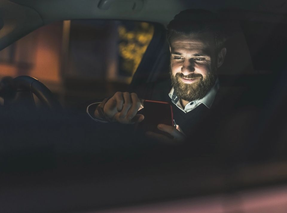Homem com barba sorrindo enquanto usa o celular dentro do carro à noite.
