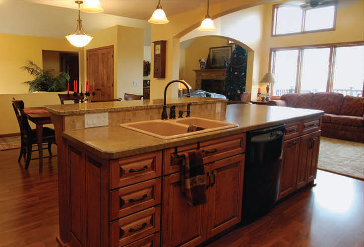 Wooden kitchen island with sink, dishwasher, cabinets, and granite countertop, with dining area in background.