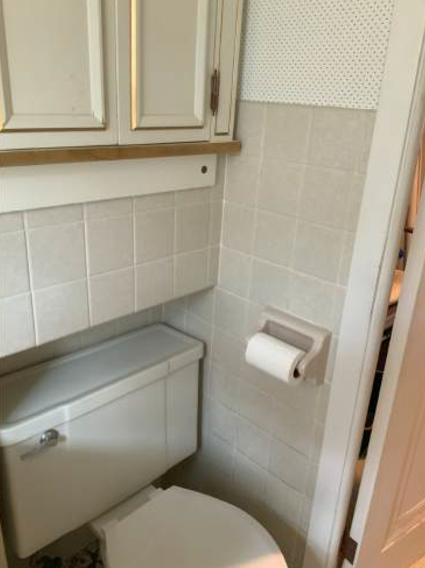 Small bathroom with white tiled walls and toilet. Toilet paper holder on the wall, and cabinets above.