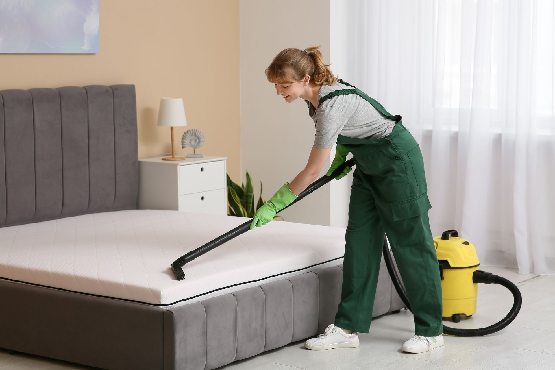 Woman in green overalls vacuuming a mattress in a bedroom with a yellow vacuum cleaner.