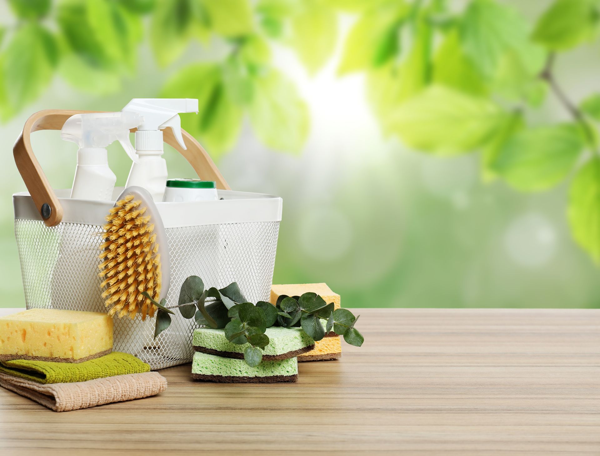 Cleaning supplies, including spray bottles, brushes, and sponges, sit on a wooden surface against a blurry green background.