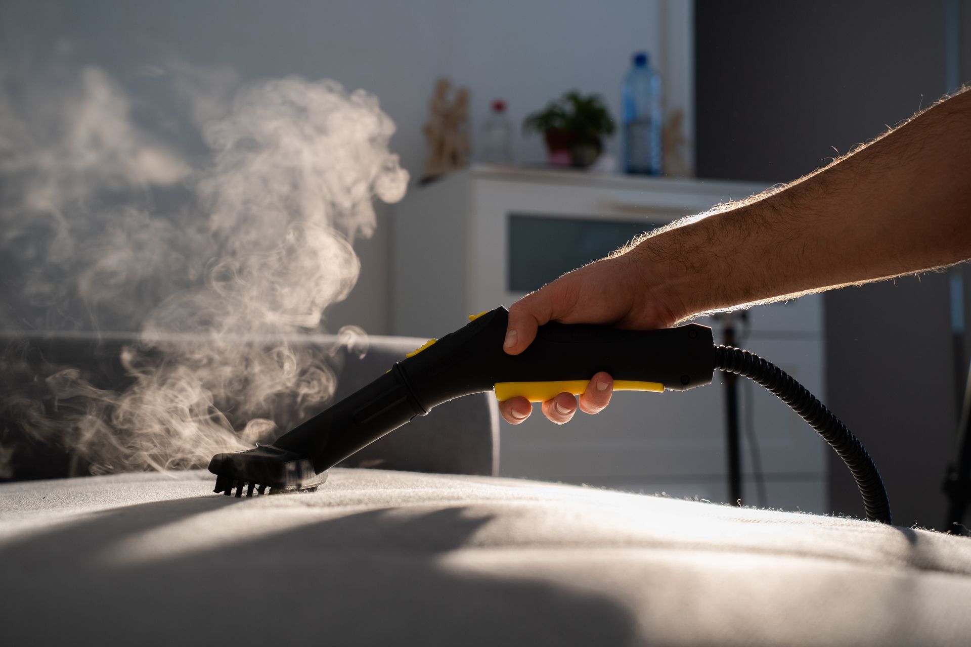 A person steam cleaning a gray couch with a black and yellow steamer, emitting white steam.