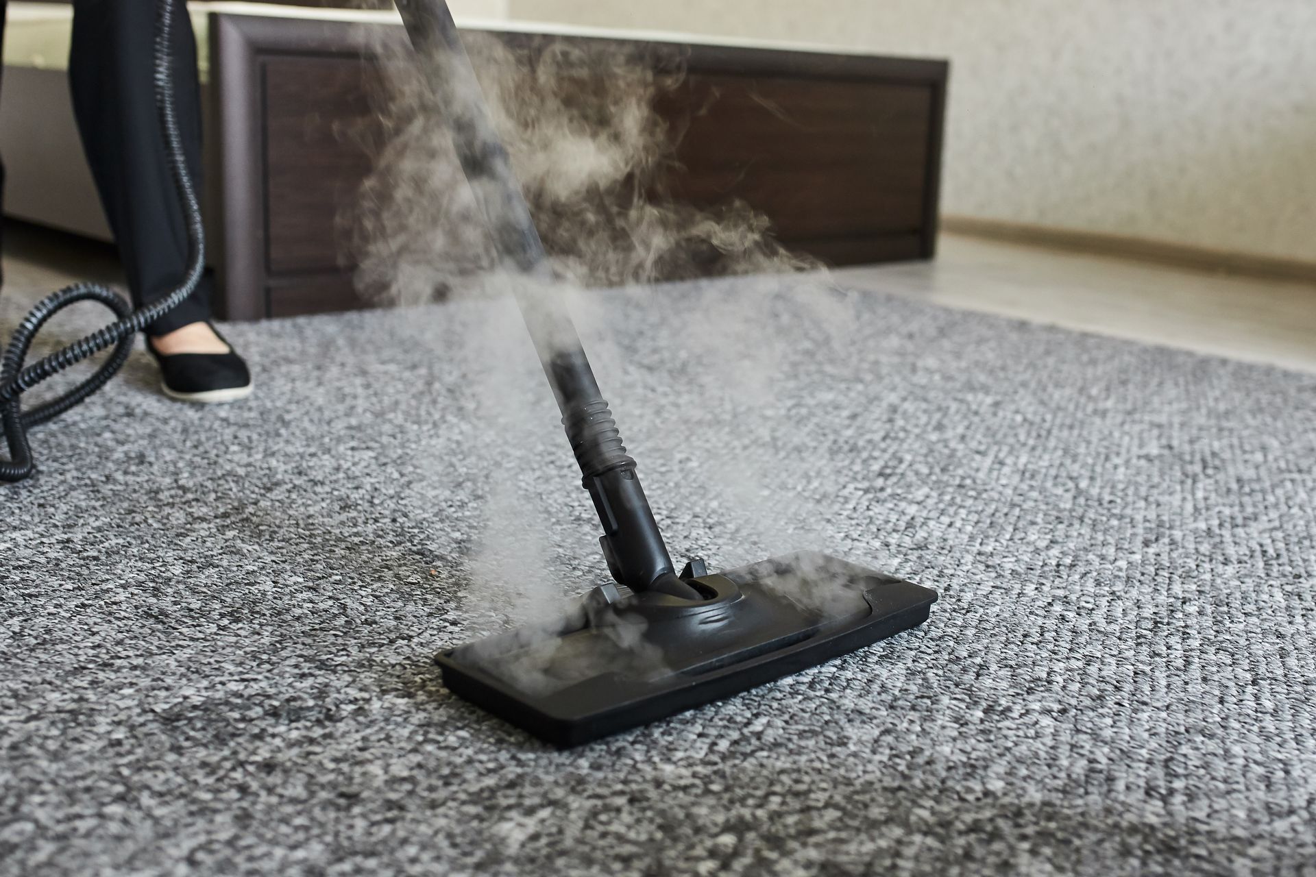 Steam cleaner on a carpet, emitting steam. A person's feet and legs are visible in the background.