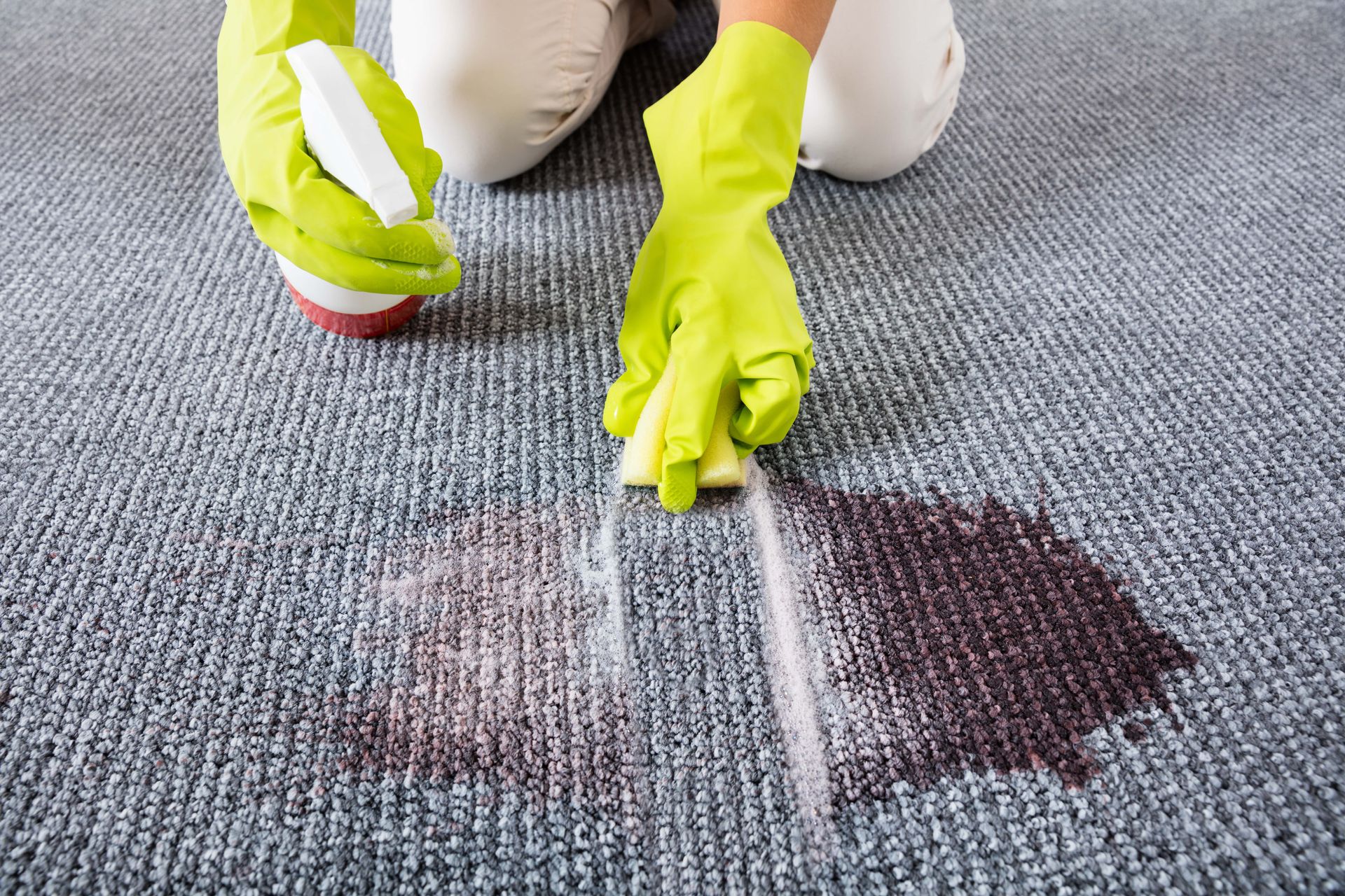Person cleaning carpet stain with spray bottle and sponge, wearing green gloves.