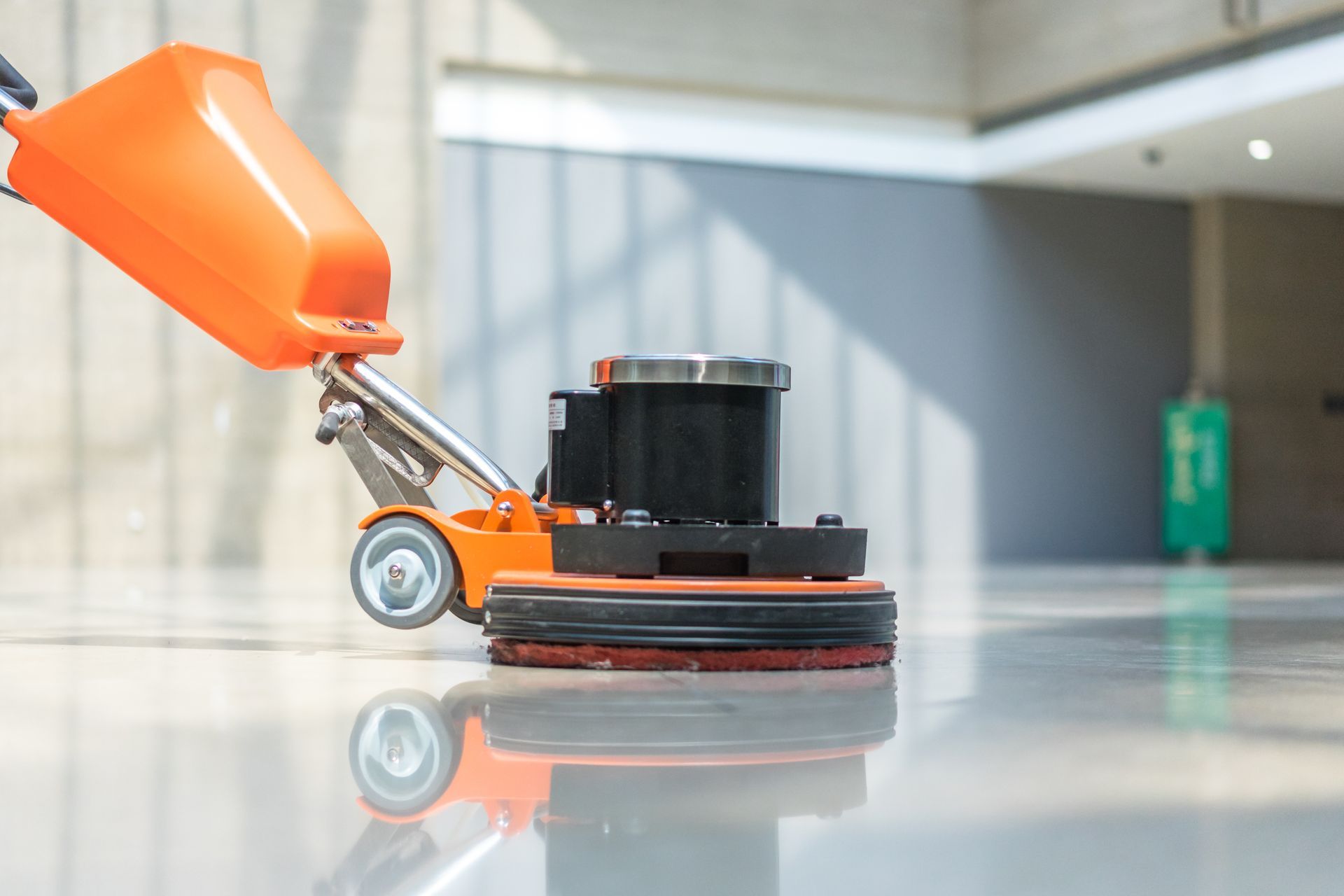 Orange floor buffer cleaning a shiny gray floor in an indoor hallway.