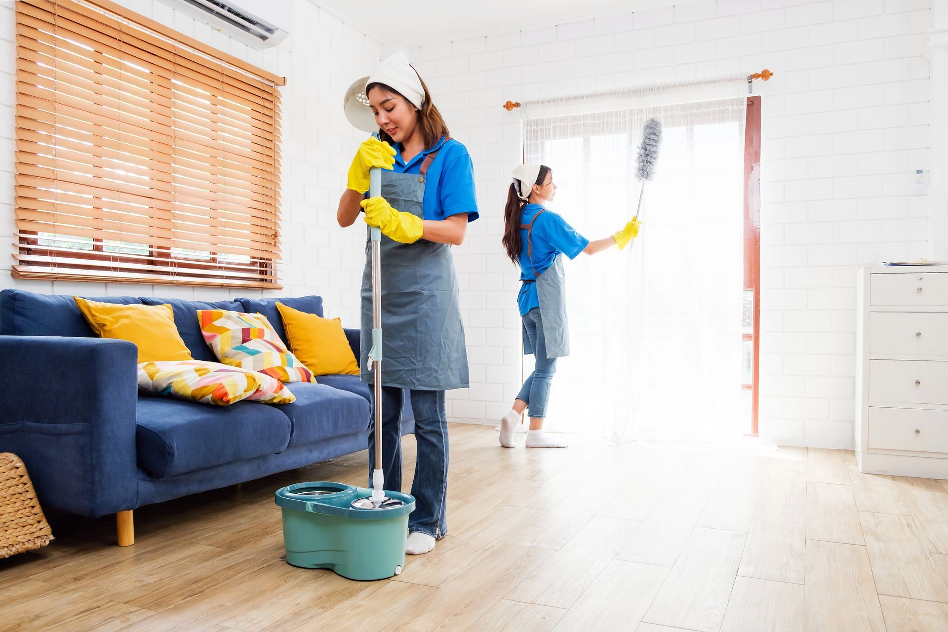 Two people cleaning a living room: one mopping, the other dusting a window.
