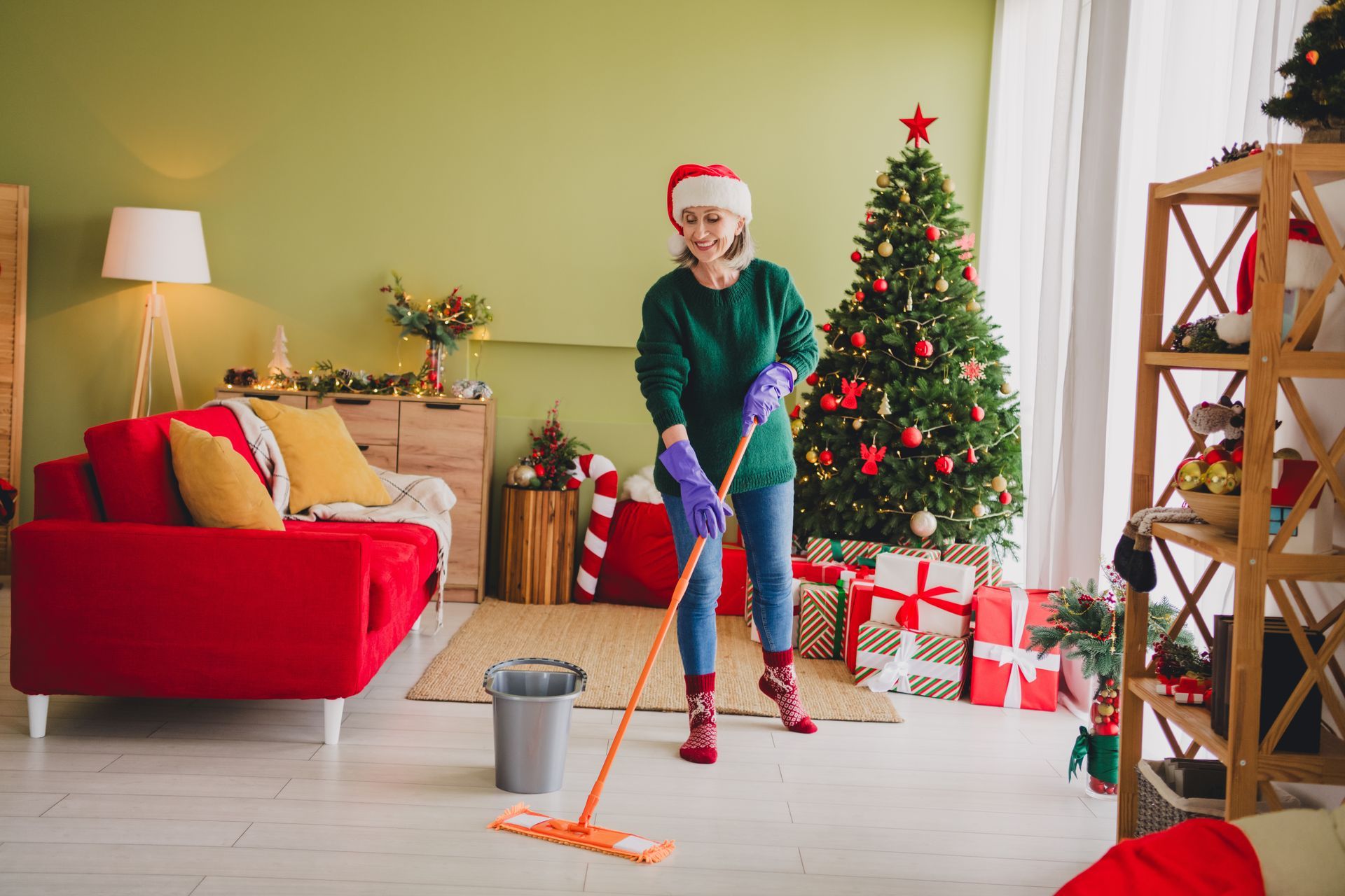 Woman in Santa hat mops a Christmas-decorated room with gifts, tree, and red couch.