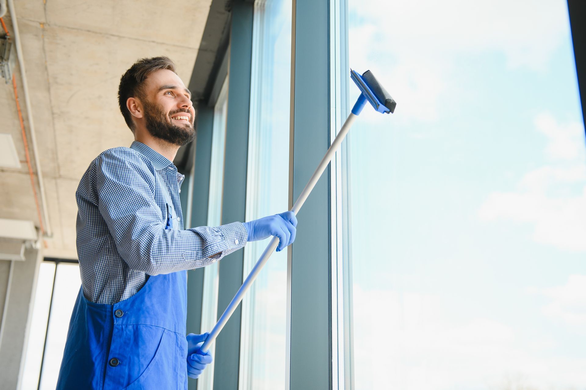 Man in blue overalls and gloves smiles while cleaning a large window with a squeegee, inside a building.
