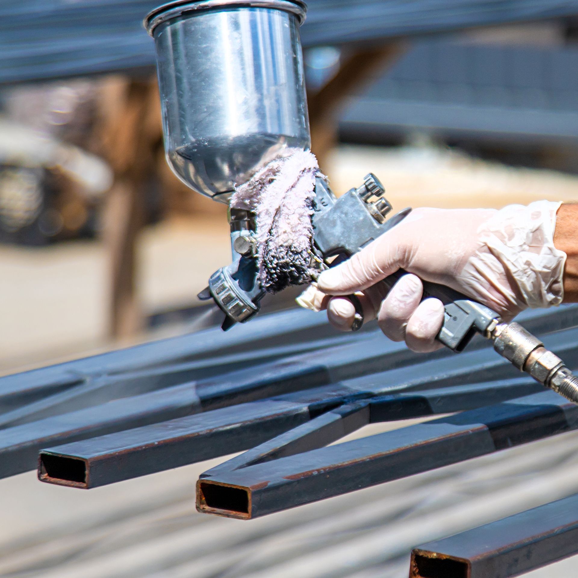 A person wearing gloves sprays dark paint onto metal bars with a silver spray gun outdoors.