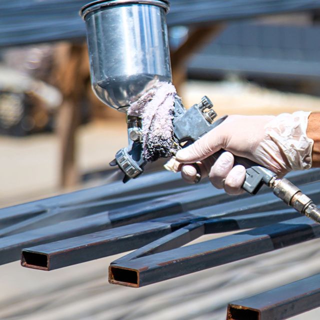 A person wearing gloves sprays paint onto metal beams using a paint gun.