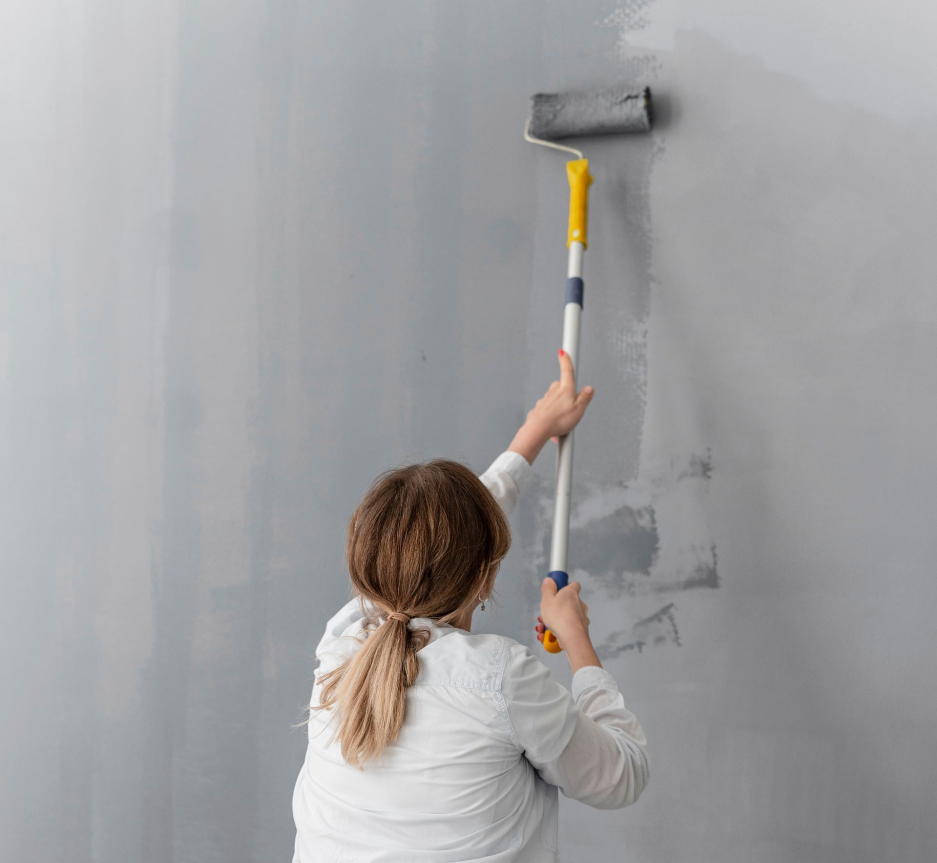 Person painting a wall gray with a paint roller, standing in a corner of a room.