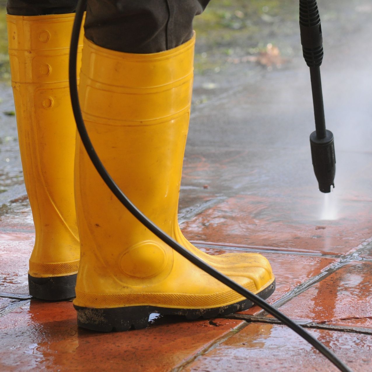 A person wearing yellow boots is using a high pressure washer to clean a sidewalk.