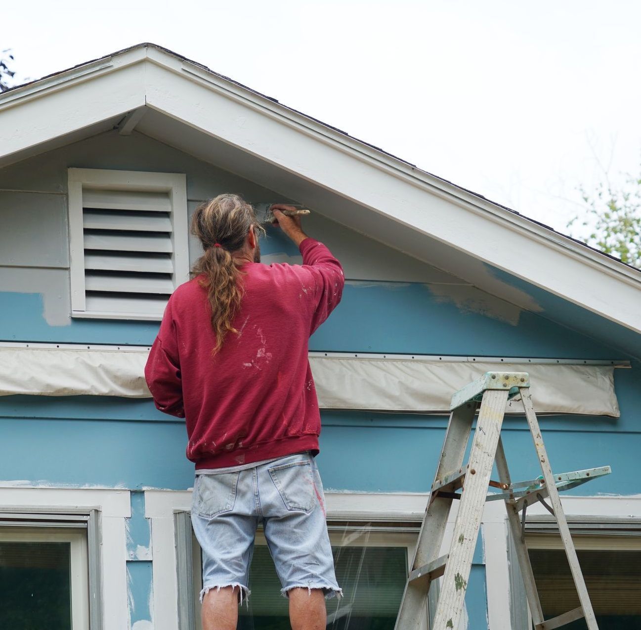 Man in red sweatshirt painting the trim of a blue house while standing on a ladder.