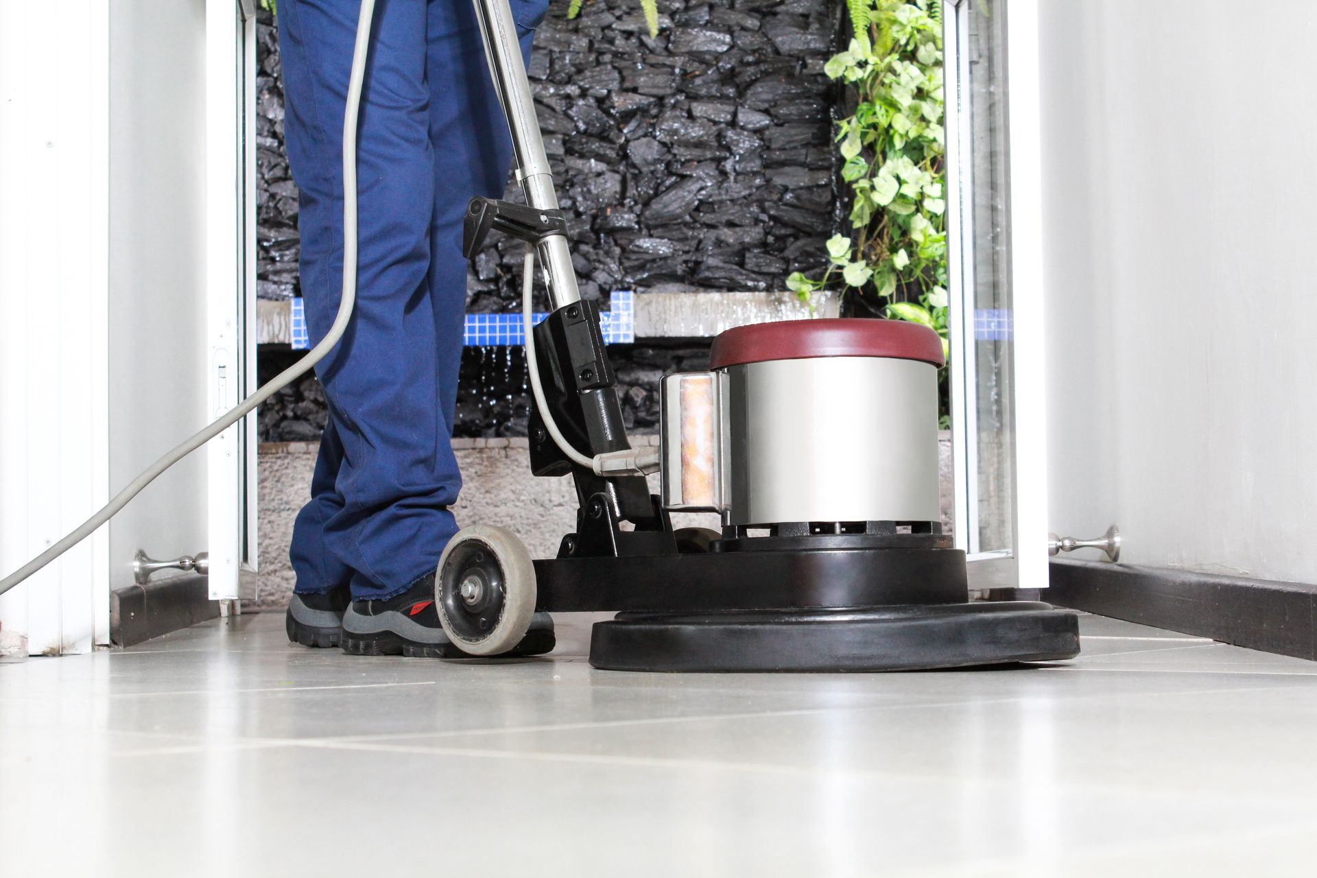 Person in blue pants using a floor buffer to clean a light-colored floor; doorway in the background.