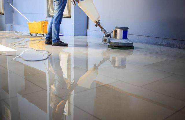 Person polishing a shiny, tiled floor with a floor machine; a yellow bucket with a mop is nearby.