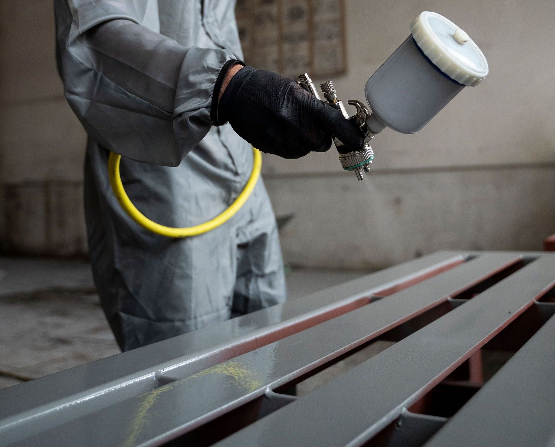Person in gray coveralls and black gloves spray paints a gray metal surface with a spray gun.
