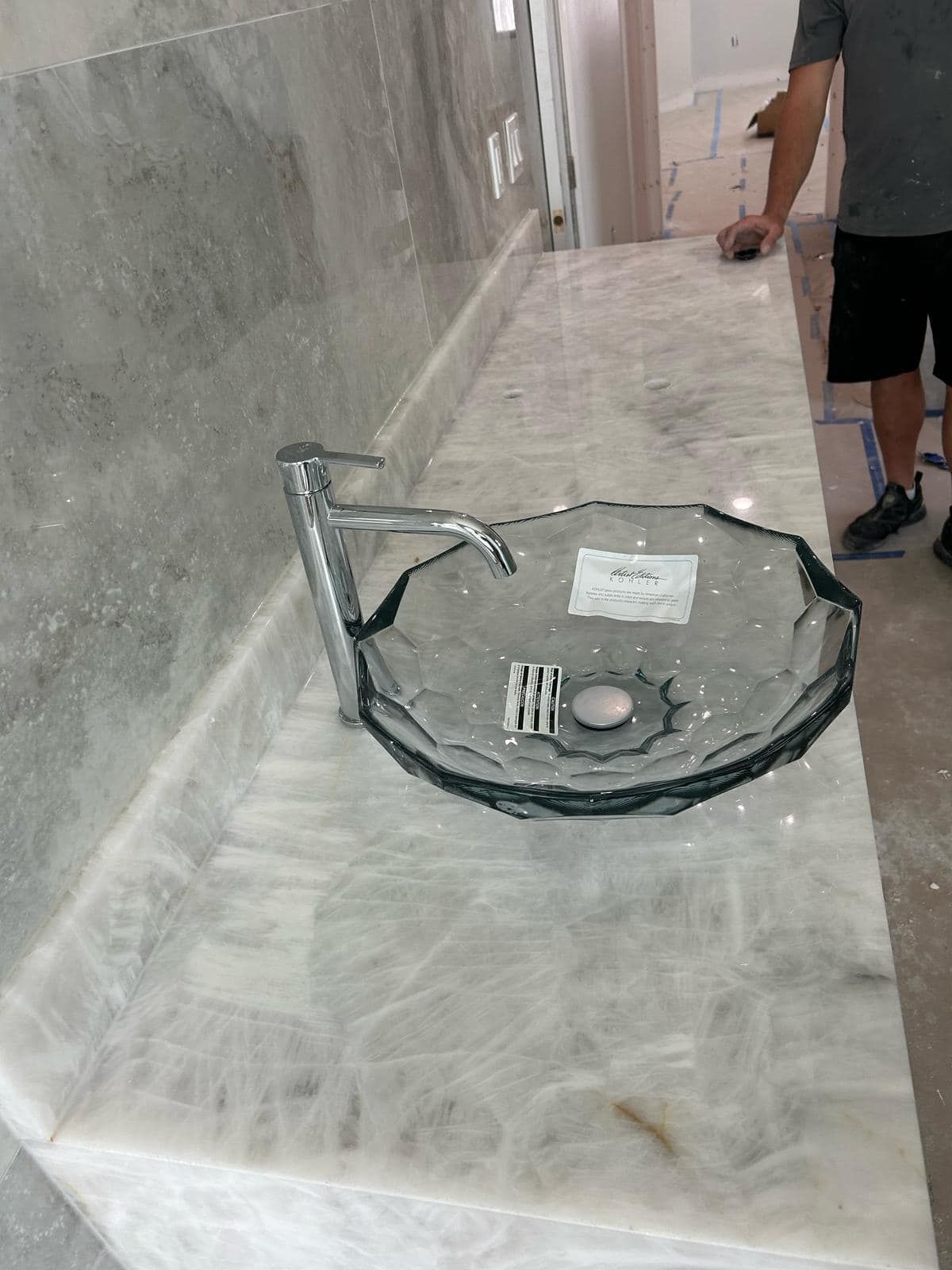 Broken glass bathroom sink on a marble countertop, with a person standing nearby.