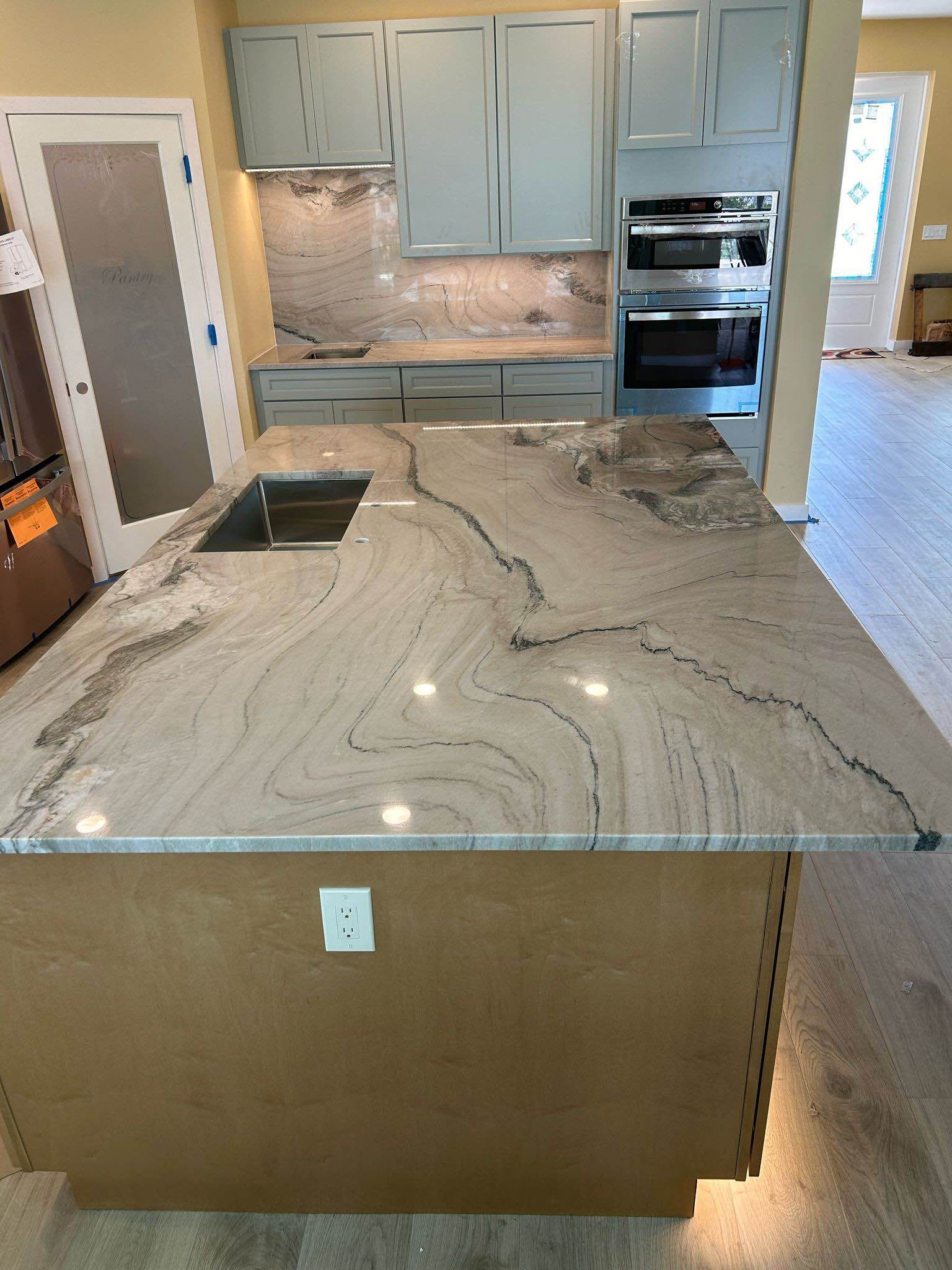Modern kitchen with large marble island, light blue cabinets, built-in oven, and pendant lighting.
