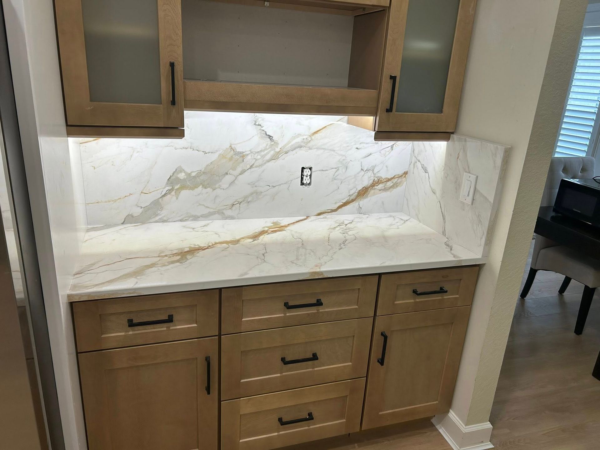 Kitchen cabinet and countertop with white marble-look backsplash and light wood cabinets under upper cabinets.