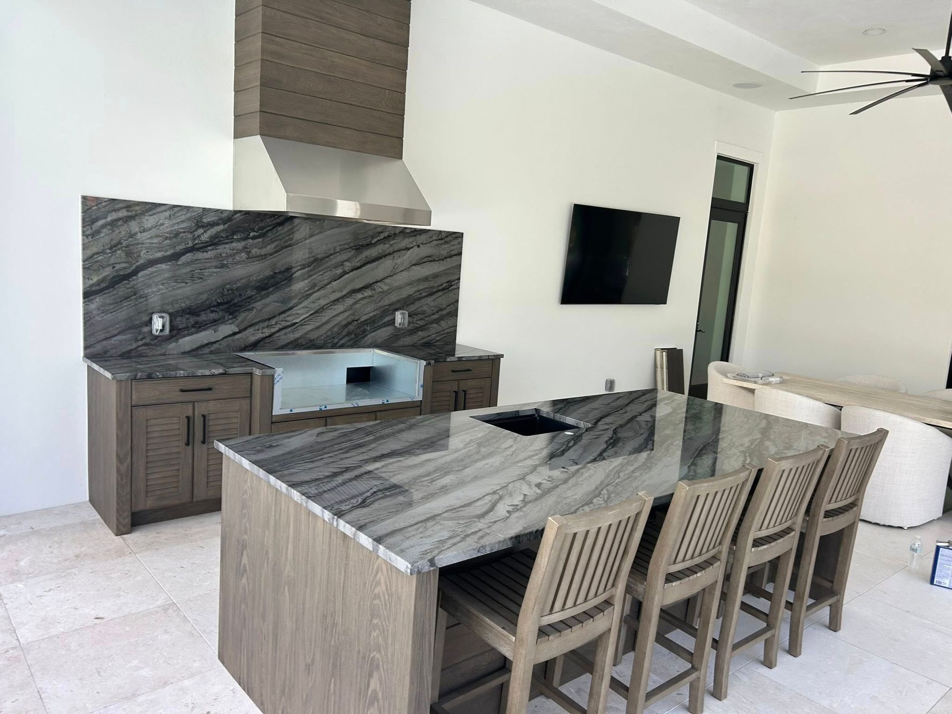 Modern kitchen with marble island, wood cabinets, and six stools beside a wall-mounted TV.