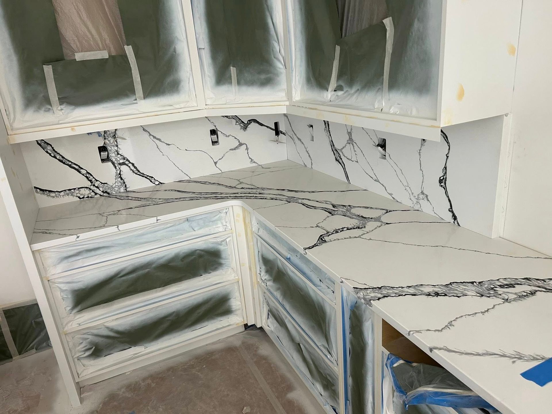 L-shaped kitchen with white cabinets and marble-look countertops under renovation, with cabinet doors removed