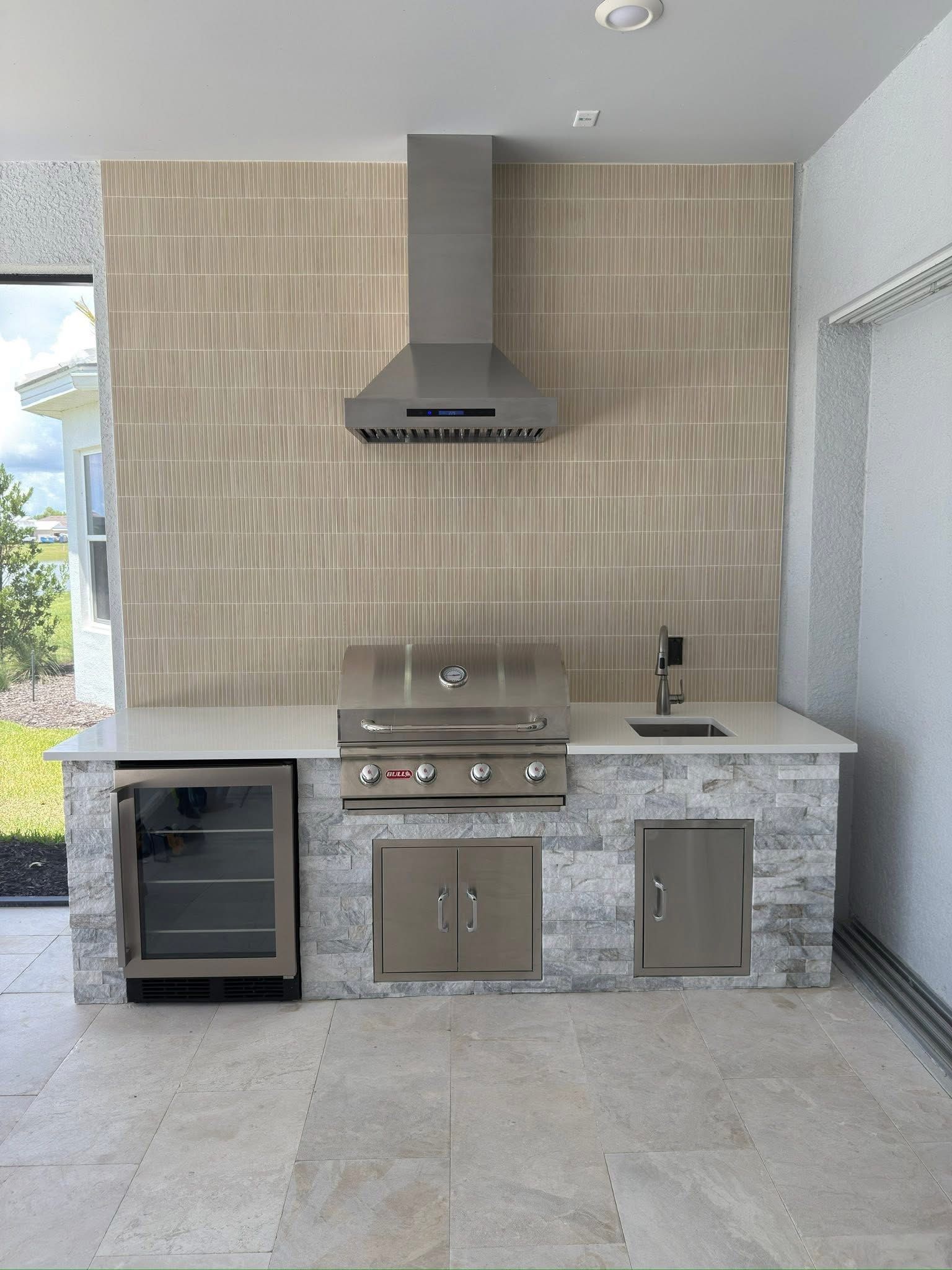 Outdoor stainless-steel grill and sink built into a stone countertop beneath a covered patio.