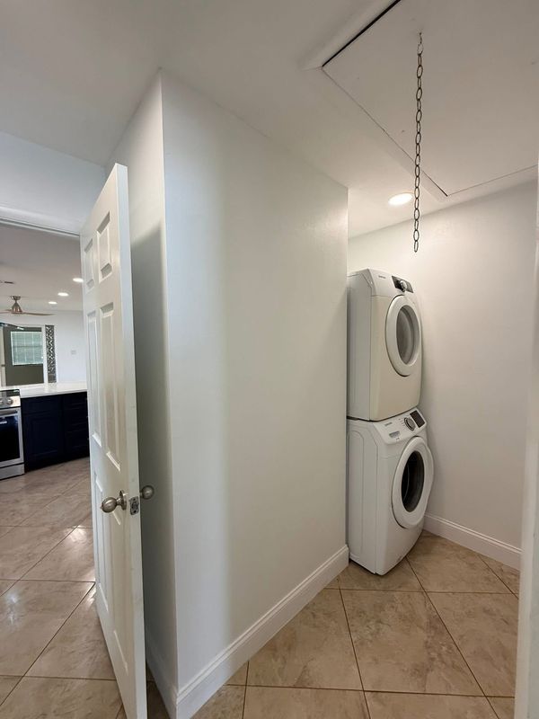 Stacked washer and dryer in a small laundry alcove with white walls and tan tile floor.
